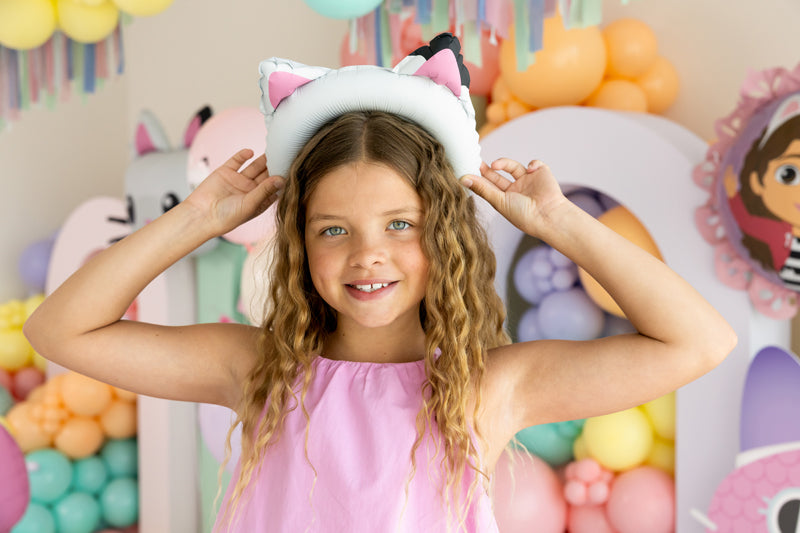 Young girl wearing a cat-themed balloon in a colorful room with balloons and decorations.