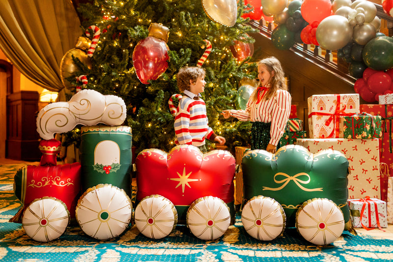 A festive Christmas-themed scene with two children standing on a large inflatable train-shaped decoration in front of a decorated tree, surrounded by wrapped gifts and balloons.